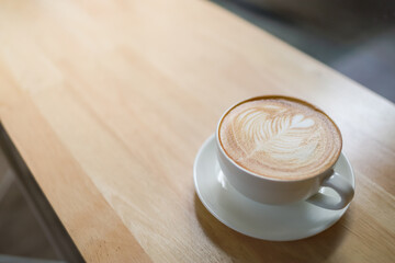 Closeup of white cup of hot coffee latte with milk foam heart shape art on wooden table under morning sunlight and shadow and copy space.