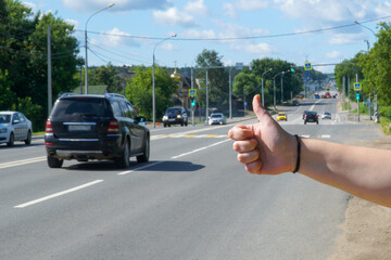   Hand of a young woman in a hitchhiking gesture against the background of a road with cars on a summer sunny day.