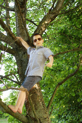 Naklejka na ściany i meble boy in dark sunglasses climbs high tree, clings to thick branches, concept of summer relaxation, dangerous outdoor activities, childish mischief