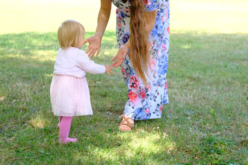 woman in a long dress, mom, teaches you to walk a little kid, a girl with blonde hair on grass in summer, concept of family summer vacation, outdoor games, first steps, child development