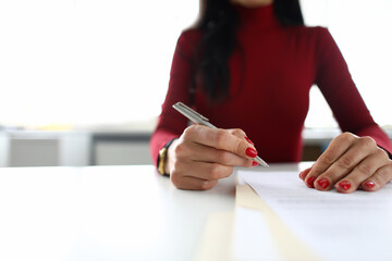 Close-up of businesswoman signing contract in modern office. Lady holding silver pen. Young woman wearing red blouse with fresh manicure. Business and career growth concept
