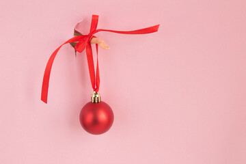 Female hand with a red christmas ball  inserted through a hole torn pink paper. Copy space. Closeup.
