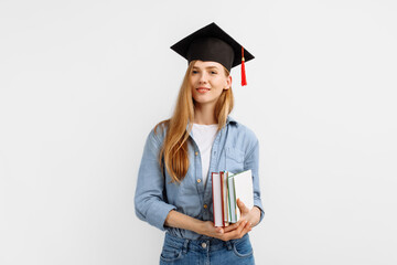 Happy beautiful girl graduate in a graduation hat on her head, holding books, standing on a white...