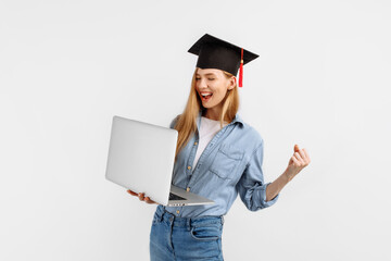 Happy excited beautiful graduate girl in a graduation cap on her head using a laptop and showing a...