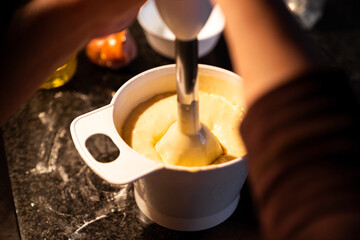 Female hands using a hand blender to mix the dough of a cake in a bowl to prepare a household...
