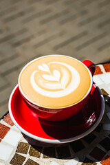 A close-up of freshly made red cup of cappuccino coffee on a mosaic stone table of a cafe terrace on a sunny day