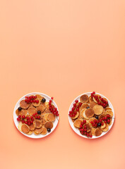Sweet mini pancakes with red and black currants on white plates on a peach background, vertical...