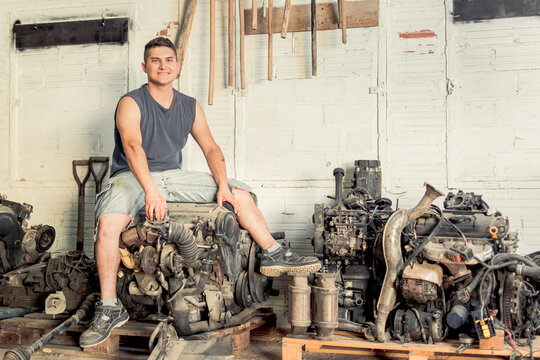 Mechanic Sitting On A Car Engine With Multiple Engines Around It.