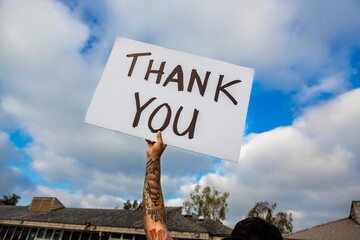 Handwritten sign stating Thank You held up by tattooed arm of man against a blue sky with clouds