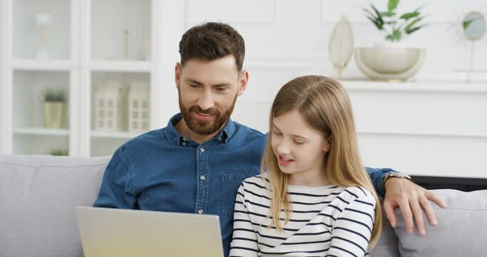 Handsome Caucasian father sitting on couch in living room and teaching small cute daughter using laptop. Little girl playing on computer with her dad and watching. video. Child spending day with daddy
