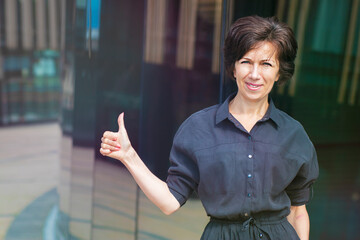 Happy positive mature adult businesswoman standing outdoors business centre showing thumb up, like...