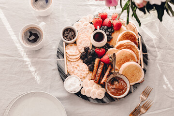 Breakfast platter. Table setting. Pancakes, breakfast sausages, berries, fruits, chocolate spread and coffee. Morning light. White and grey colors. Peonies. Summer time. Sunday brunch party. Weekend