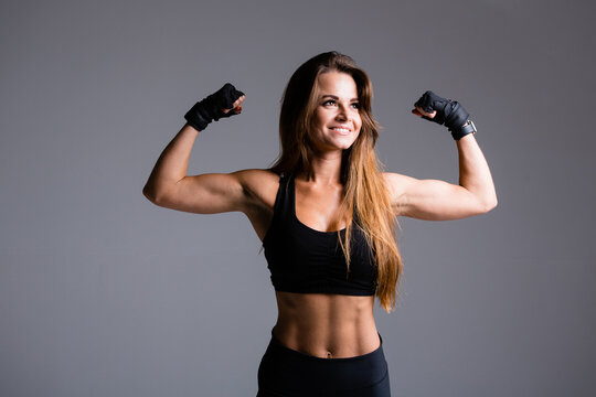 Beautiful boxer woman with long haired shows arm muscles, biceps and triceps on a gray background in gloves in a black sports suit © DmitryStock