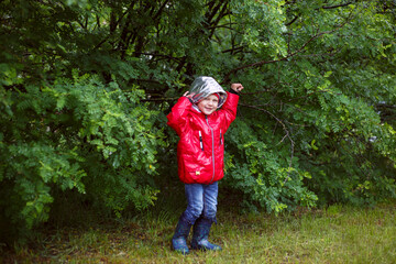 a boy in a red jacket with a hood hides from the rain under an acacia bush, holds a branch with his hand and laughs.
lmage with selective focus