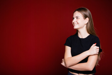 Fototapeta premium A European girl in a black T-shirt looks away and hugs herself on a red background.