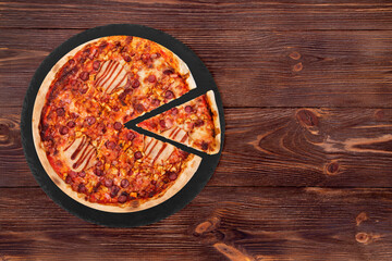 BBQ pizza with ham, bbq sauce, bacon and salami, with a slice slightly removed on slate black platter which is on wooden table, top view and space for text