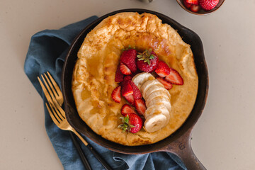 Dutch baby pancake baked until golden brown in cast iron skillet and topped with strawberries, banana and maple syrup. Weekend brunch meal. Sunday breakfast. Simple recipe. Fluffy battered pancake.