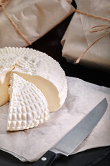 Adyghe cheese cut off a triangular piece. Nearby are three cheese heads packed in paper. On a wooden board. Background gray concrete