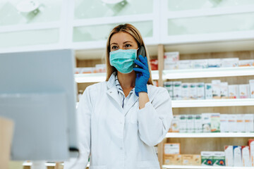 Female pharmacist with protective mask on her face talking on phone while working at pharmacy....