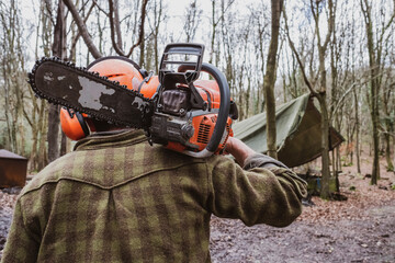 Rear view of man wearing safety gear walking through forest carrying chainsaw on his shoulder.