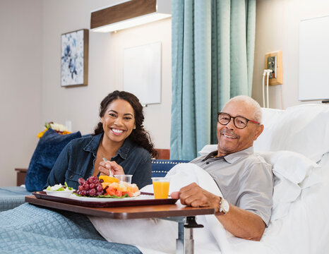 Daughter Visiting Senior Father Eating Healthy Meal In Bed In Nursing Home