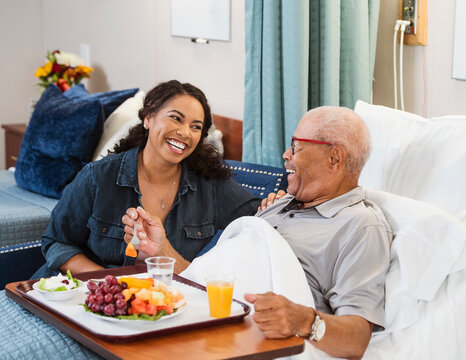 Daughter Visiting Senior Father Eating Healthy Meal In Bed In Nursing Home