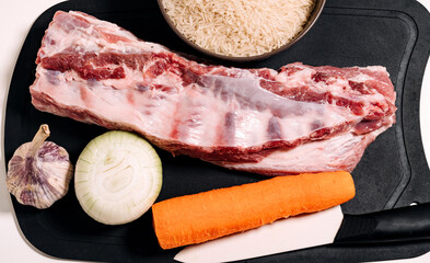 Uncooked long grain rice in a deep bowl. In the background is a piece of meat, an onion, carrots and garlic.