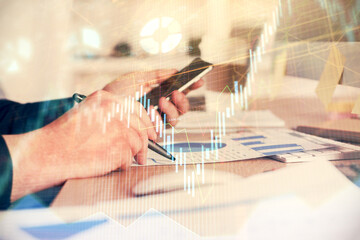 Double exposure of man's hands holding and using a phone and financial graph drawing. Analysis concept.