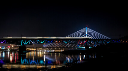 Night time panorama of modern european bridge. Blurred lights from passing by train. Iconic colourful bridge in Warsaw, Poland.