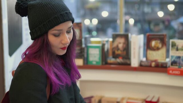Millenials lifestyle. Portrait of stunning asian woman with bright red lips and purple hair reading a book in a bookstore. teenage student shopping. Concept of hipster style and trends.