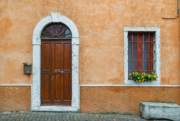 Exterior of an old building with an arched door and a window with a flowering potted plant on an orange wall, Italy