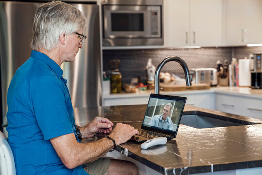 Senior man video conferencing with doctor on laptop computer with contactless thermometer on counter next to him in kitchen