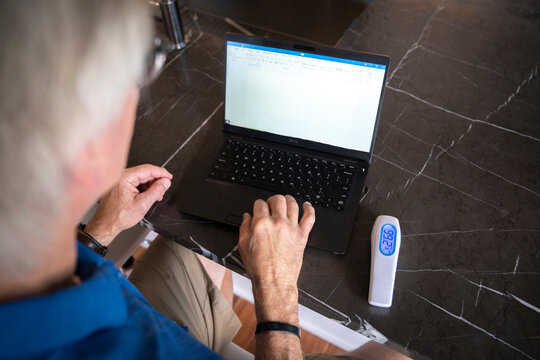 Close up of mature man's hands on keyboard of laptop with contactless thermometer next to him