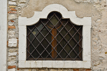 Close-up of the shaped window of an old building with an iron grate on a weathered brick wall, Italy