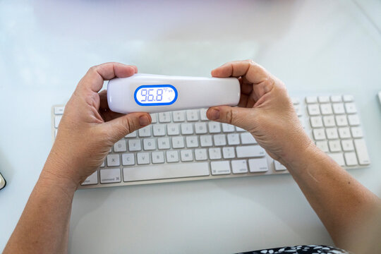 Close up of woman's hands holding contactless digital thermometer over computer keyboard