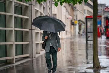 Middle-aged businessman wearing a suit caught out in the rain during a windy  drizzly day fighting the wind under an umbrella in Holborn, London during the COVID-19 pandemic 101