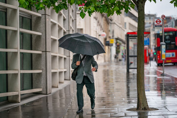 Middle-aged businessman wearing a suit caught out in the rain during a windy  drizzly day fighting the wind under an umbrella in Holborn, London during the COVID-19 pandemic 100