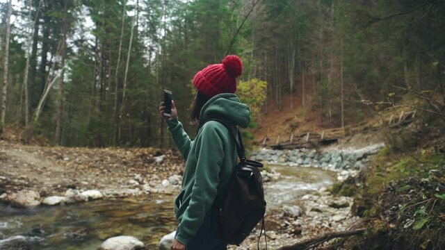 Woman in hike wear with backpack take panorama shots on smartphone or mobile phone while standing on shore of mountain river. Millennial using modern technology to making or taking photo for memory.