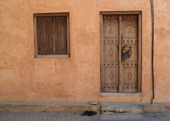 A pair of sandals in front of a wooden door closed with a golden padlock in a traditional building in Al Ain, United Arab Emirates. The window is also made by wood and the wall is terracotta colored.