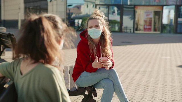 Two familiar young girls greet each other by waving at hand at a safe social distance. They communicate in the distance in medical masks. Concept of pandemic, coronavirus, epidemic, quarantine.