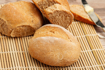 close up image of home made whole wheat bread loaves fresh out of oven. They are cooling on a bamboo mat on wooden table. A butter knife with butter on it is there to put delicious spread on a slice.