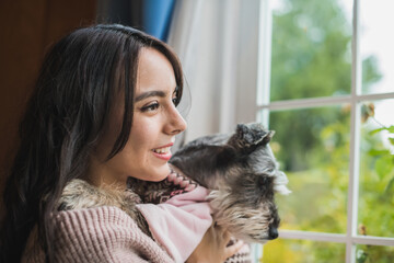 Girl looking out the window carrying her pet in her arms, dressed in a pink sweater, her dog Schnauzer enjoys the company of her friend, an afternoon of relaxation and company of her pet. 2