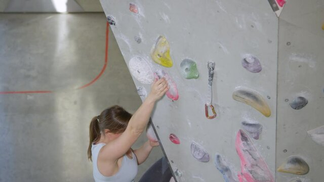 SLOW MOTION, CLOSE UP: Strong woman reaches up to grip a crimp hold while lead climbing in a cool indoor training center. Woman loops her belay rope into a carabiner while holding on to a crimp hold
