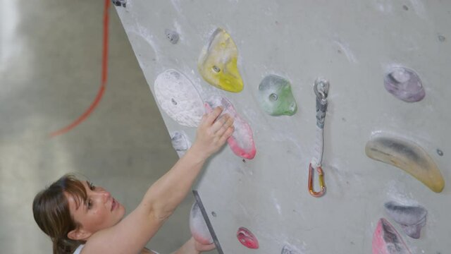 SLOW MOTION, CLOSE UP: Woman loops her belay rope into a carabiner while holding on to a crimp hold. Strong woman reaches up to grip a crimp hold while lead climbing in a cool indoor training center.