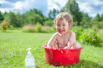 Little baby washes in red basin on the green grass. Little baby having fun, splash water and laughs. Happy child bathing outdoors on green grass in spring garden. Summer, vacation, health