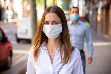 Young woman in disposable face mask walking along city street. Concept of Covid 19 virus spread prevention and human safety.