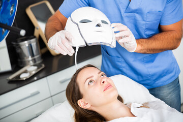 Man cosmetologist putting on LED mask preparing woman for skin rejuvenation therapy in aesthetic...