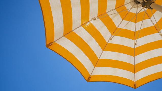 White and yellow striped beach umbrella. Blue sky in the background. View from below. Relaxing context. Summer holidays by the sea. General contest and location