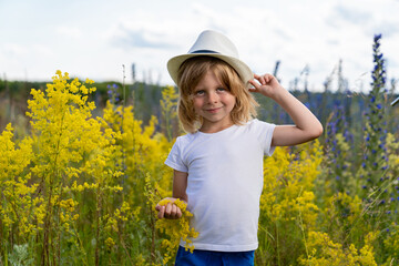 a boy with blond hair in a straw hat stands in a field with beautiful wild flowers