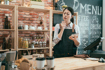 Portrait of young barista woman standing behind counter thoughtfully looking on wooden table at cafe bar. pensive female waitress in apron and white shirt in coffee shop holding note frowning face.